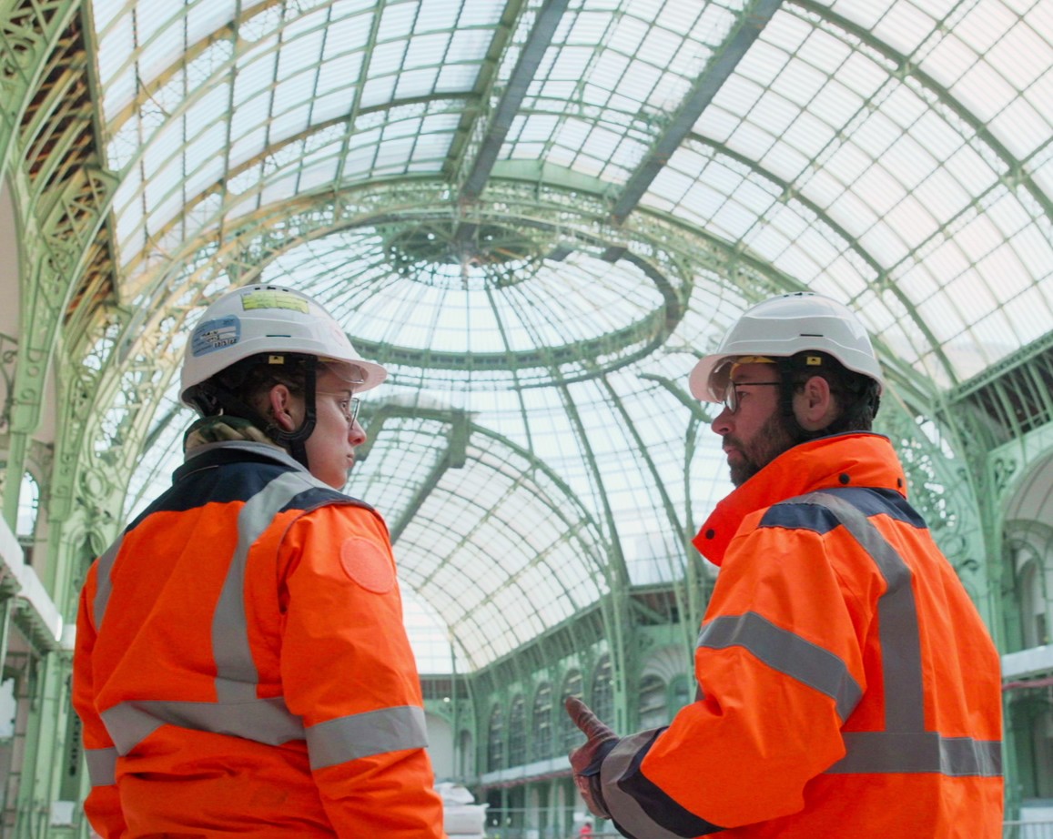 Two workers in orange safety jackets and white hard hats discussing a glass-roofted building